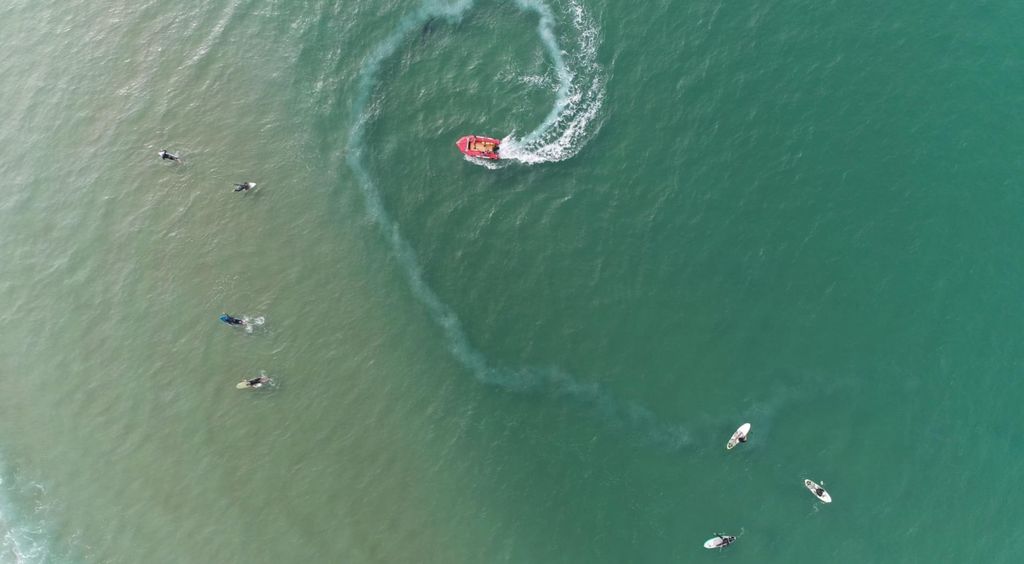 Slika mora iz zraka sa surferima i brodom te morski pas koji izvire iz vode