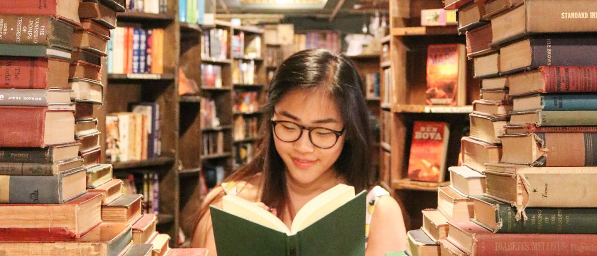 žena s naočalama sjedi i čita knjigu u knjižnici okružena drugim knjigama