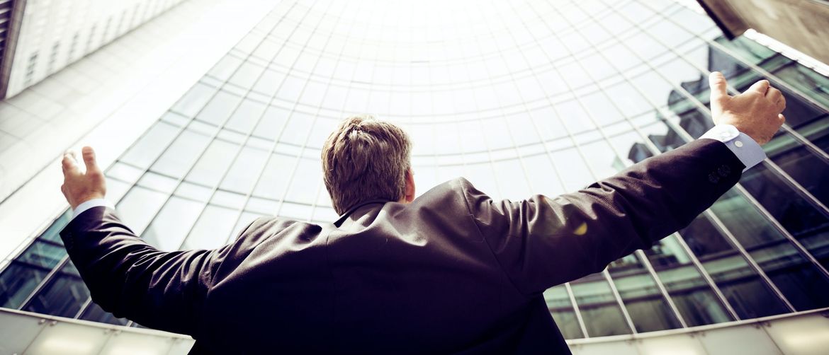 muškarac stoji ispred poslovnog nebodera i šire ruke u zrak kao znak uspjeha
