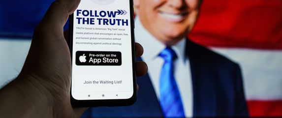 Mobitel u rukama s opisom aplikacije Truth Social, slika Trumpa u pozadini