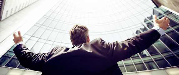 muškarac stoji ispred poslovnog nebodera i šire ruke u zrak kao znak uspjeha