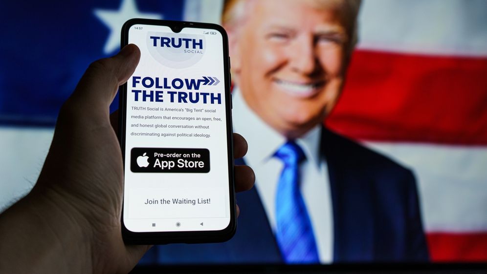 Mobitel u rukama s opisom aplikacije Truth Social, slika Trumpa u pozadini